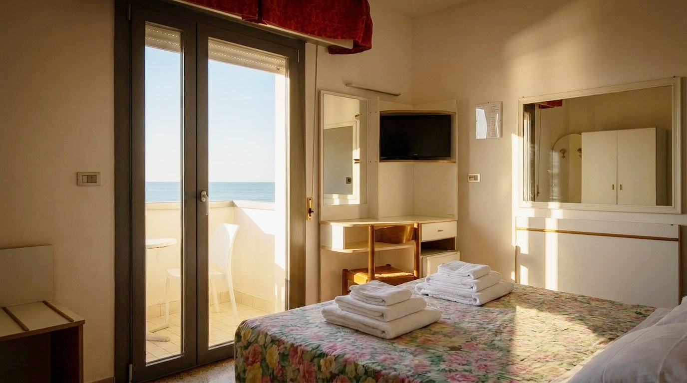 Camera vista mare dell'Hotel Columbia — porta-finestra sul balcone con l'Adriatico sullo sfondo, letto matrimoniale e luce dorata del mattino